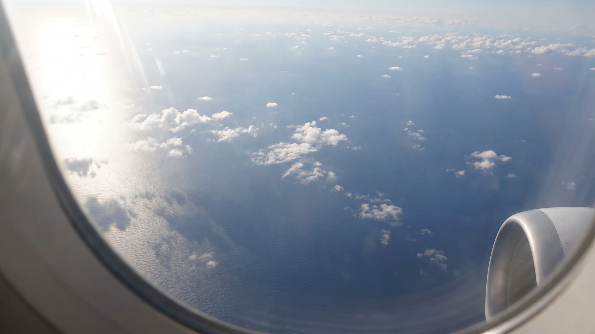 Thin, broken low clouds over a dark blue ocean, seen from an aircraft window